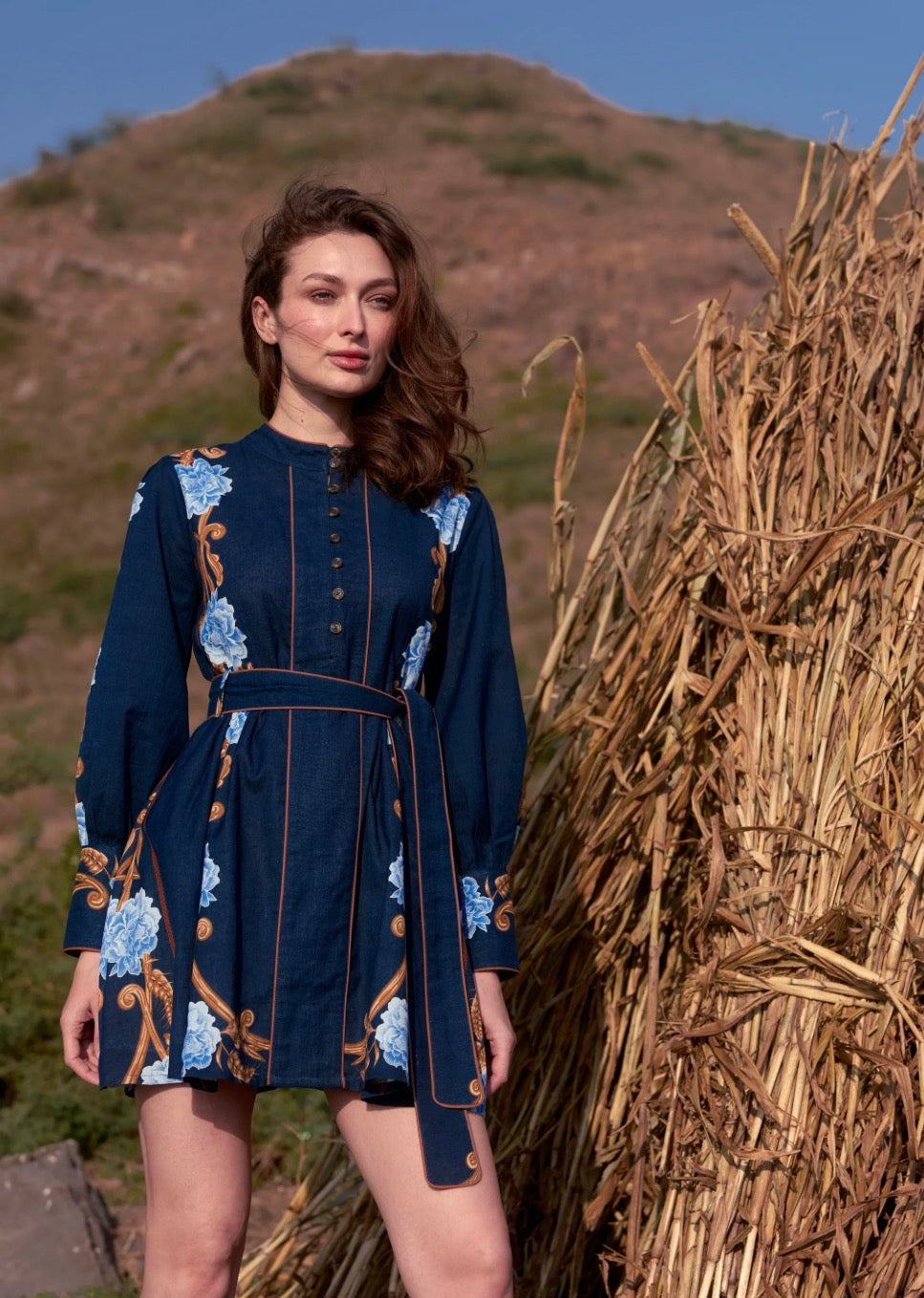 A woman wearing a navy blue long-sleeve mini dress with blue and white floral patterns and gold trim, standing outdoors next to a large bundle of dried grass with a hill in the background.