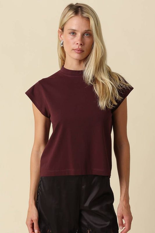 A blonde woman wearing a short-sleeve crewneck top in deep burgundy, paired with black pants, standing against a solid beige background.