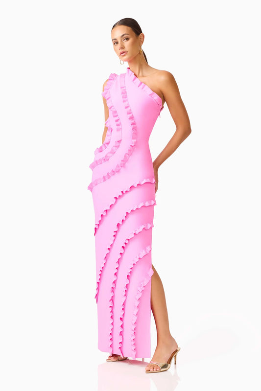 A woman modeling a bright pink one-shoulder maxi dress with cascading ruffle details and a high side slit, paired with metallic heeled sandals, against a plain white studio background.