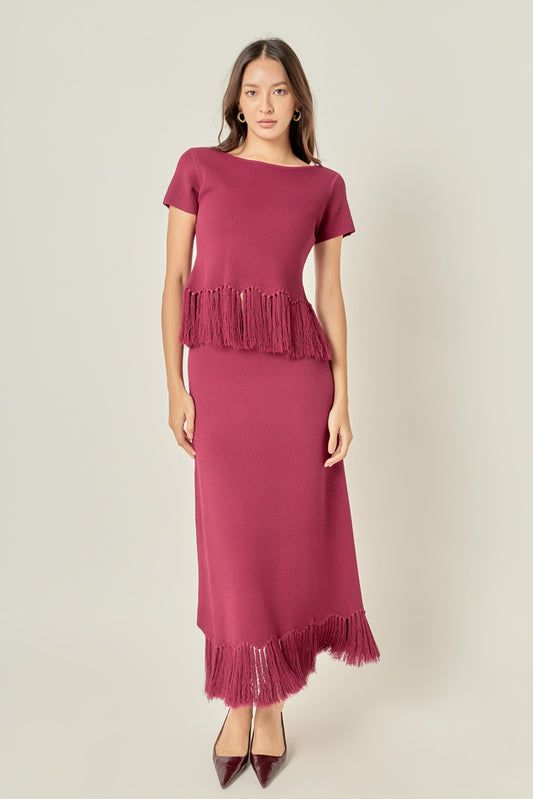 A woman modeling a dusty rose pink knit dress with short sleeves, a boat neckline, and fringe detailing at the peplum and hem, paired with dark brown pointed-toe heels against a light gray studio background.