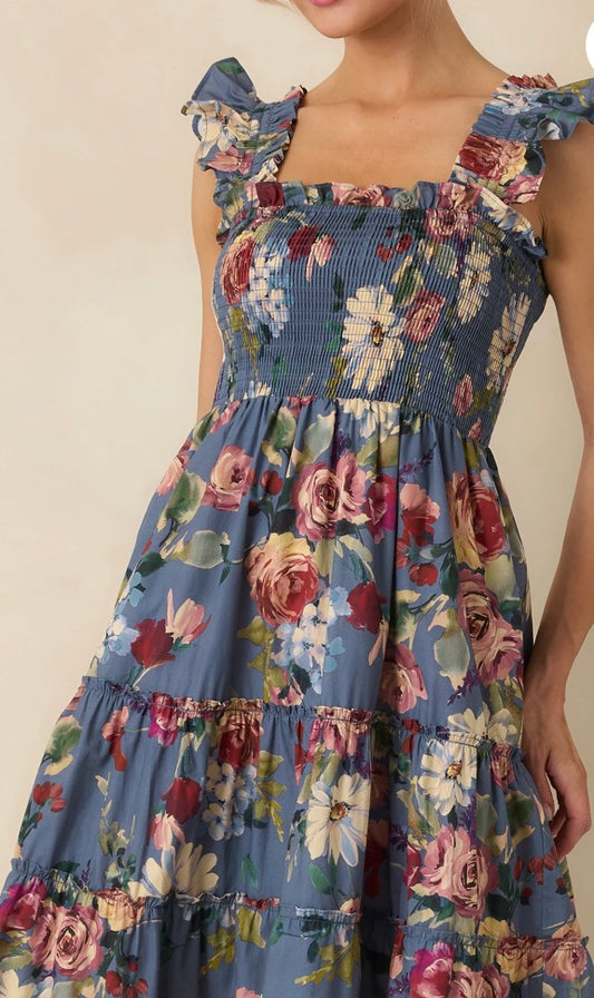 A close-up of a woman wearing a tiered blue floral dress with ruffled shoulder straps and a smocked bodice, shown against a plain beige background.