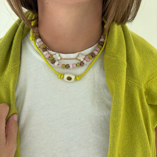 A close-up of a person wearing layered beaded necklaces. The most prominent necklace is a bright yellow cord with a central white oval bead featuring a green eye motif.