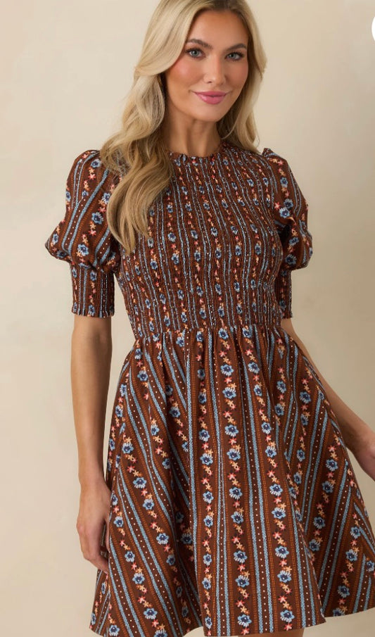 A woman modeling a brown smocked dress with vertical blue and orange floral stripes, set against a plain beige background.