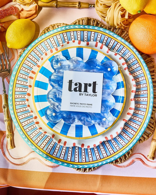 A decorative round magnetic photo frame with a blue and white striped center and a turquoise rim, styled on a table with lemons, pasta, and gold-toned flatware. The center card reads 'tart BY TAYLOR'.