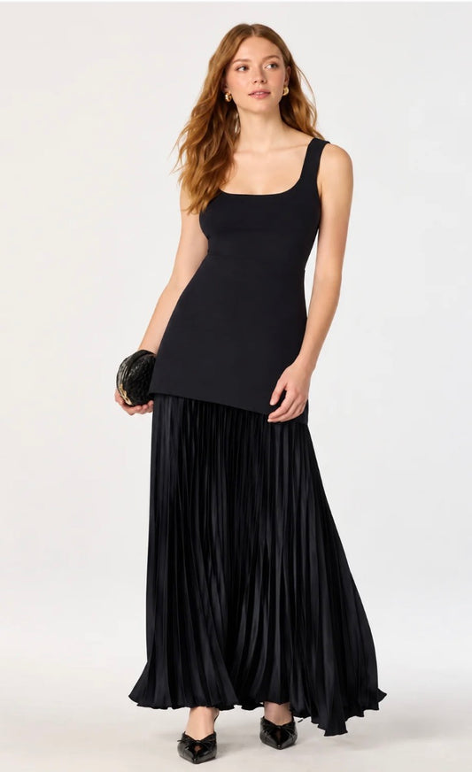 A woman modeling a black sleeveless maxi dress with a square neckline and a long, pleated skirt, holding a small black clutch against a plain white studio background.