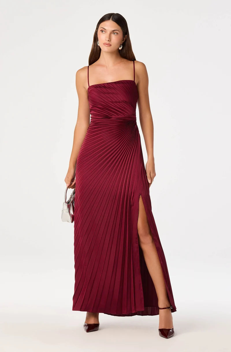 A woman modeling a floor-length pleated maxi dress in deep burgundy with thin spaghetti straps, a draped bodice, and a high thigh slit, photographed against a clean light gray studio background.