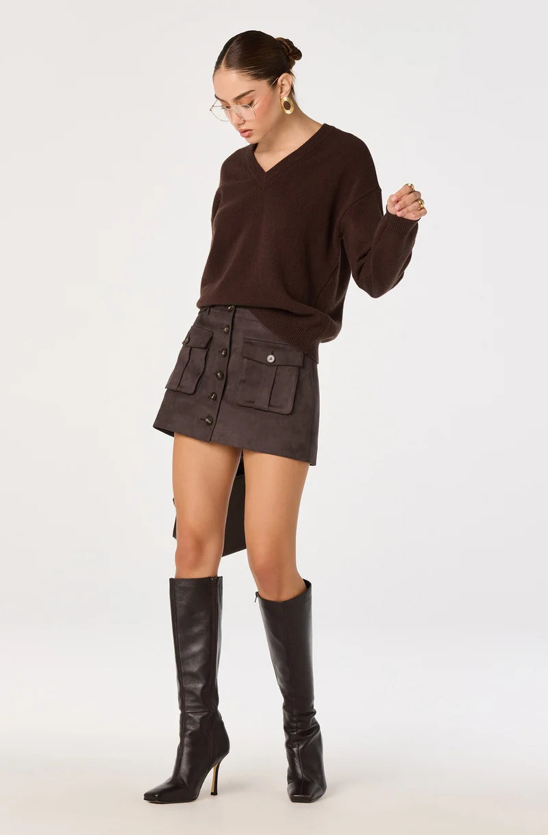 A model wearing a dark brown long-sleeve knit sweater, a dark brown button-up mini skirt with utility pockets, and black knee-high leather boots, standing against a light gray studio background.