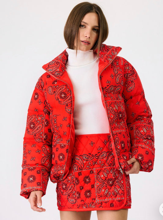 A model wearing a red puffer jacket and matching red mini skirt with a black paisley bandana print, styled over a white mock neck top, against a plain white background.
