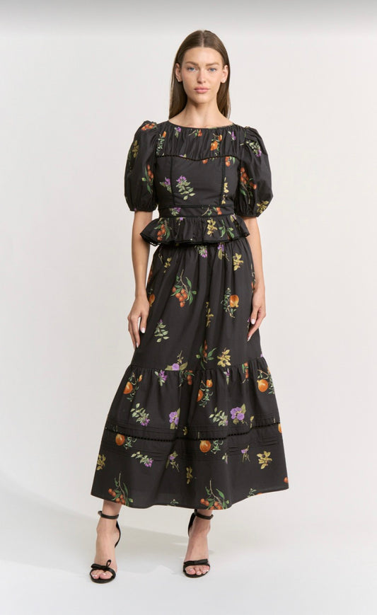 A model wearing a black midi dress with a colorful floral and fruit print, featuring puff sleeves and a ruffled waist, paired with black strappy heels against a light gray studio background.