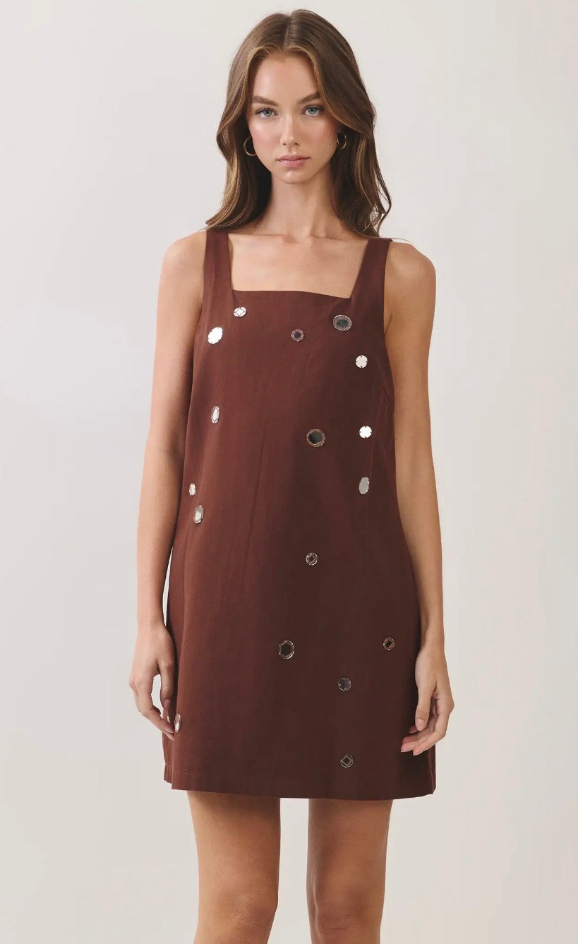 A woman modeling a sleeveless brown mini dress with a square neckline and scattered silver circular embellishments, set against a plain light gray studio background.