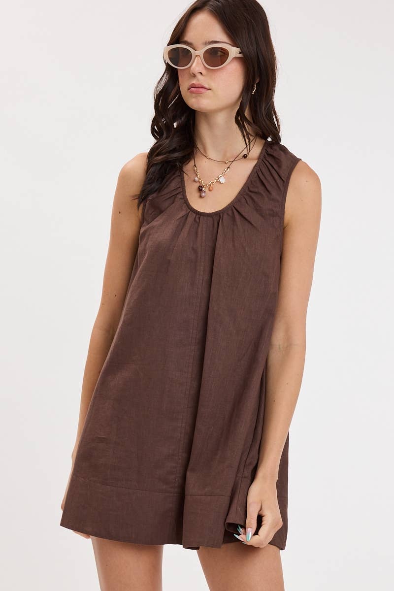 A woman modeling a sleeveless brown linen dress with a gathered neckline and a loose, flowy silhouette, set against a plain white studio background.