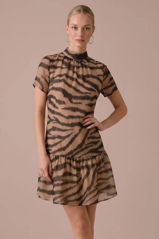 A woman modeling a short-sleeve high-neck mini dress with a brown and beige zebra print against a solid warm beige studio background.