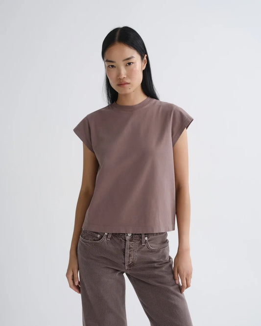 A woman modeling a muted mauve-brown short-sleeve crop top with a relaxed fit, paired with dark brown pants, against a plain light gray studio background.