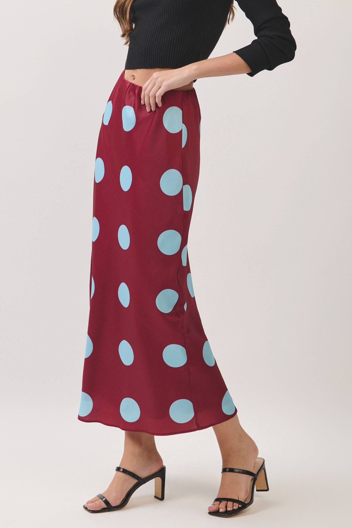 A model wearing a long maroon maxi skirt with large light blue polka dots, paired with a black ribbed top and black strappy high-heeled sandals, against a light gray studio background.