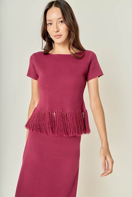 A woman modeling a short-sleeve knit top and matching midi skirt in a deep berry pink. The top features a textured waffle knit body and a fringed peplum hem.