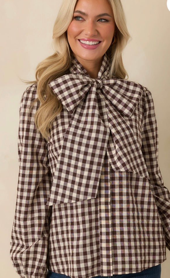 A smiling blonde woman wearing a brown and beige gingham long-sleeve blouse with a large bow at the collar, photographed against a plain light beige background.