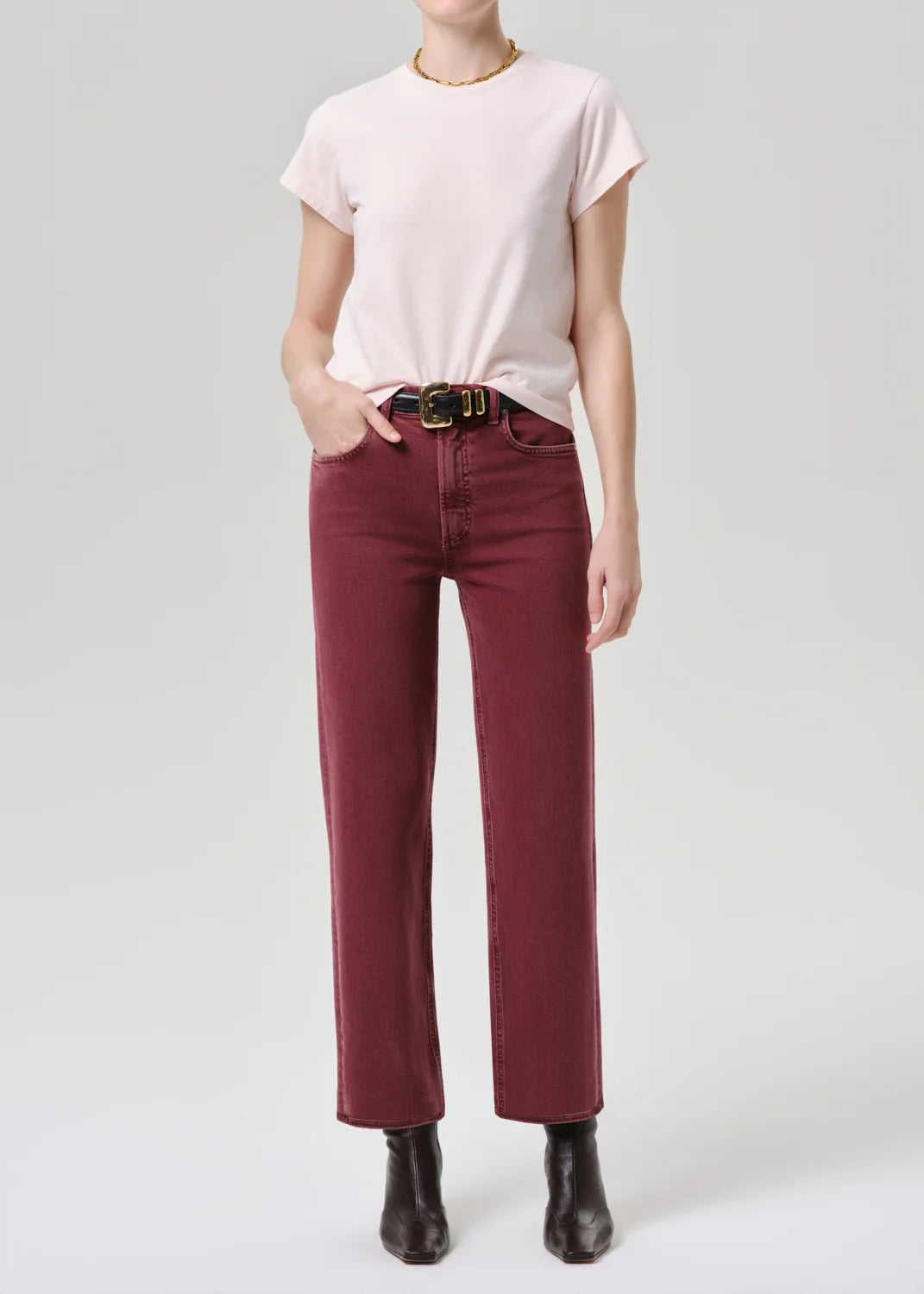 A model wearing a light pink short-sleeve t-shirt, dark red wide-leg jeans with a black belt, and black ankle boots, standing against a light gray studio background.