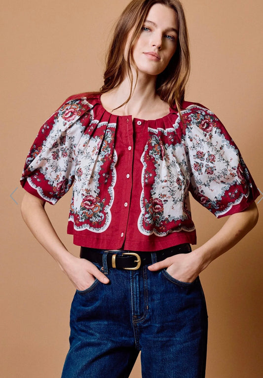 A woman modeling a cropped short-sleeve blouse with a red and white floral pattern, paired with dark blue jeans and a black belt, against a solid tan studio background.