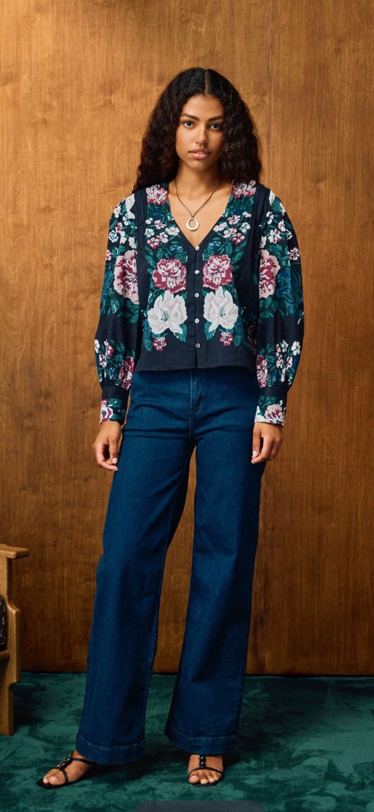 A woman stands against a wood-paneled wall wearing a dark floral cardigan with blue wide-leg jeans and brown sandals.