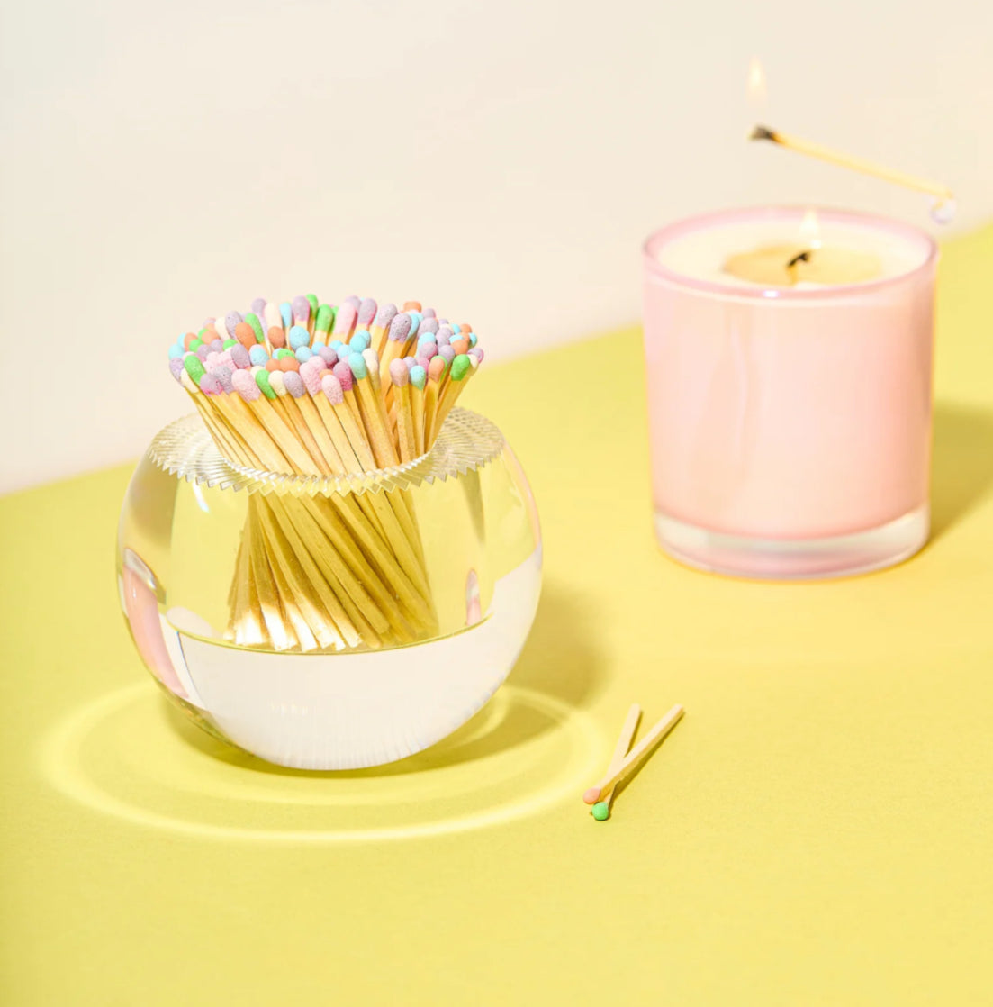 Glass container with colorful matches and a pink candle on a yellow surface