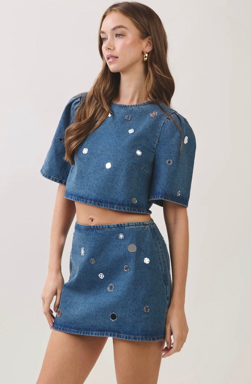 A model wearing a blue denim two-piece set consisting of a short-sleeve cropped top and a matching mini skirt, both embellished with silver metallic studs, against a plain light background.
