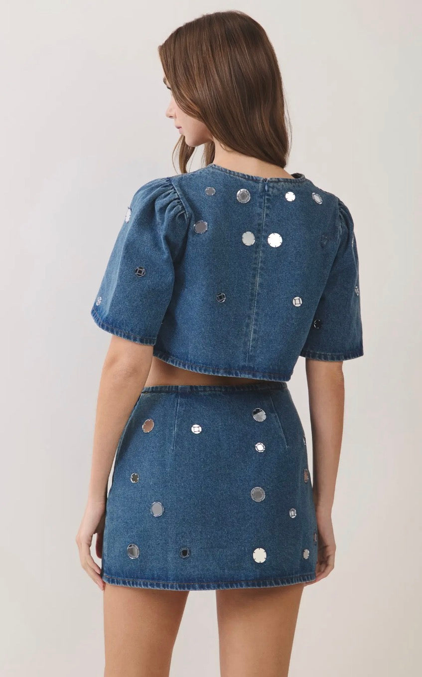 A model shown from the back wearing a blue denim two-piece set consisting of a short-sleeve crop top and a matching mini skirt, both decorated with scattered silver circular embellishments, against a plain light background.