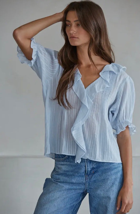 A woman wearing a light blue and white vertically striped ruffled blouse with puff sleeves, paired with blue jeans, photographed against a neutral light gray background.