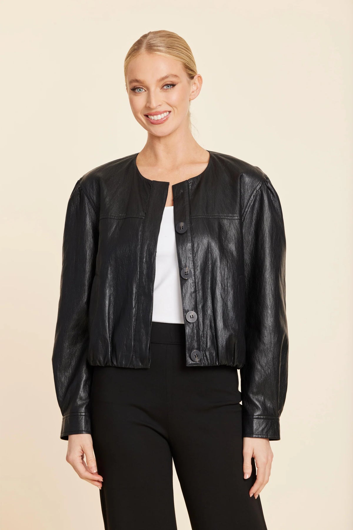 A woman modeling a cropped black faux-leather jacket with a button-front closure over a white top and black pants, photographed against a plain beige studio background.
