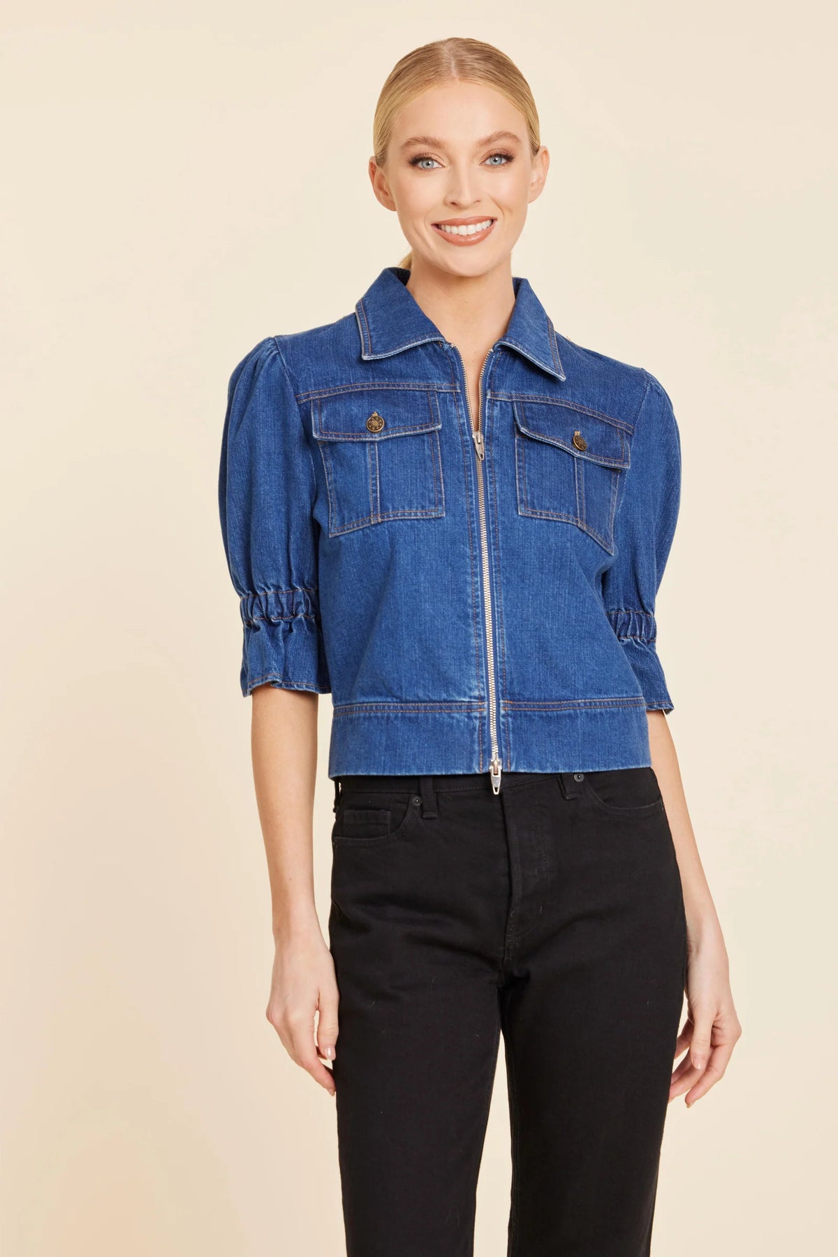 A woman modeling a cropped blue denim jacket with puff sleeves, a front zipper, and chest flap pockets, paired with black pants against a plain beige studio background.