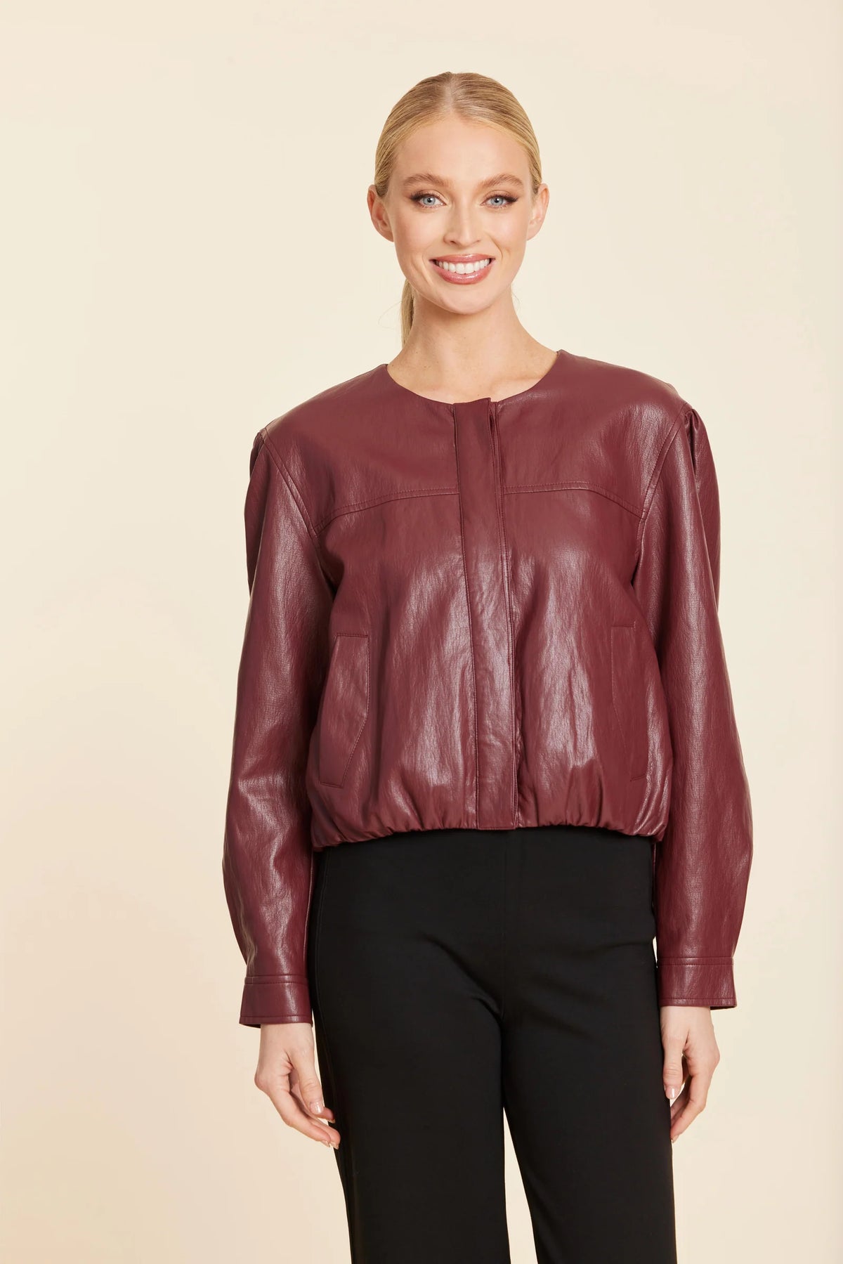 A woman modeling a cropped, long-sleeve burgundy faux-leather jacket with a gathered waist, paired with black pants, against a solid beige background.
