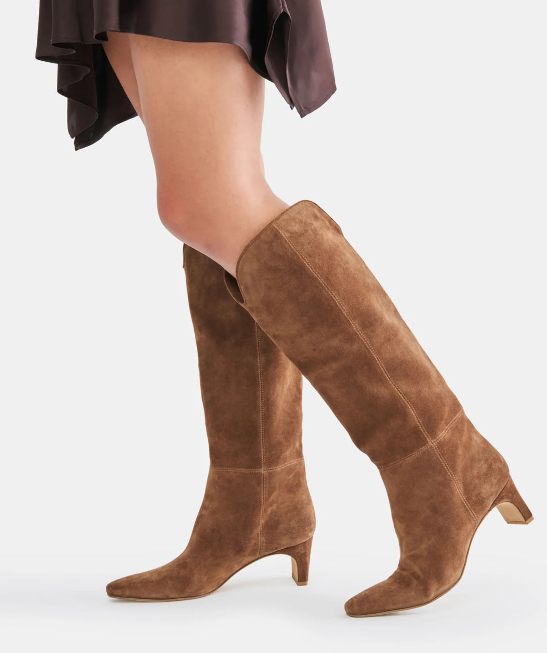 A pair of knee-high brown suede boots with a pointed toe and low block heel, shown on a model's legs against a light gray studio background.
