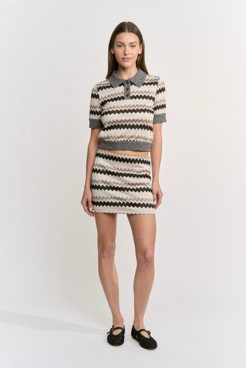 A model stands against a plain light gray studio background wearing a matching knit two-piece set consisting of a short-sleeve cropped polo-style top and a high-waisted mini skirt. The fabric has a repeating chevron-like horizontal pattern in off-white, black, and gray, with a solid gray collar and cuffs.