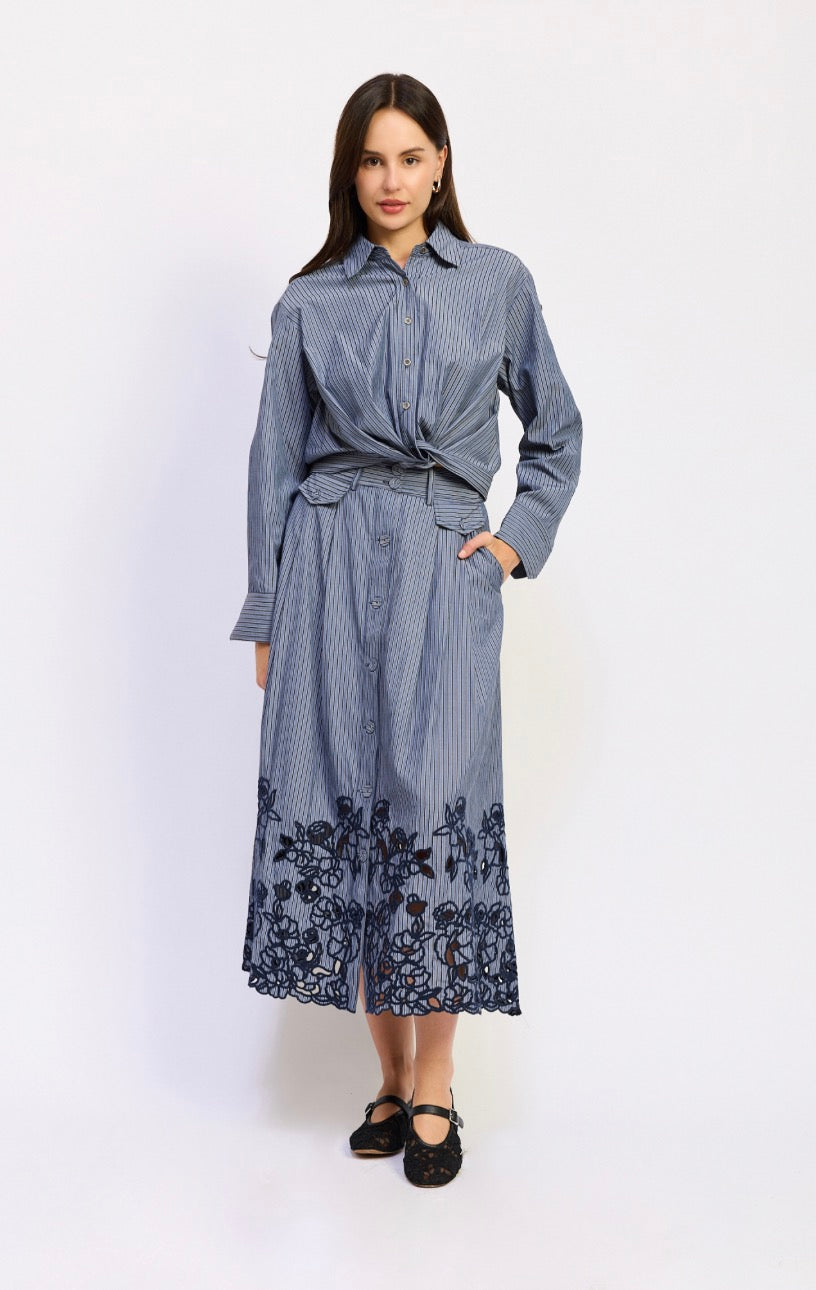 A woman models a long-sleeve, button-up midi dress with a blue and white vertical stripe pattern, a matching tie belt, and dark floral lace embroidery along the hem, paired with black flats against a plain white studio background.