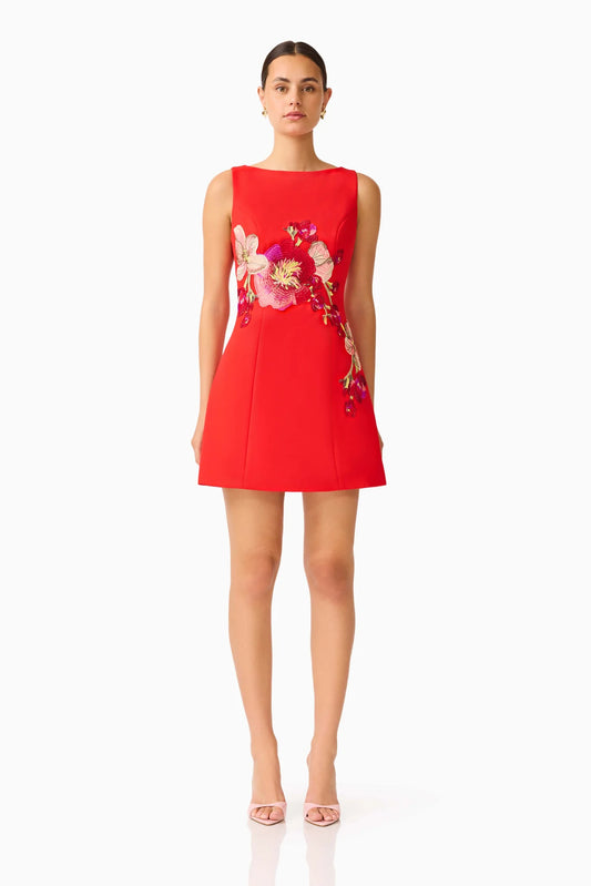 A woman modeling a sleeveless red mini dress with a large floral print on the bodice and upper skirt, paired with clear high-heeled sandals, against a plain white studio background.