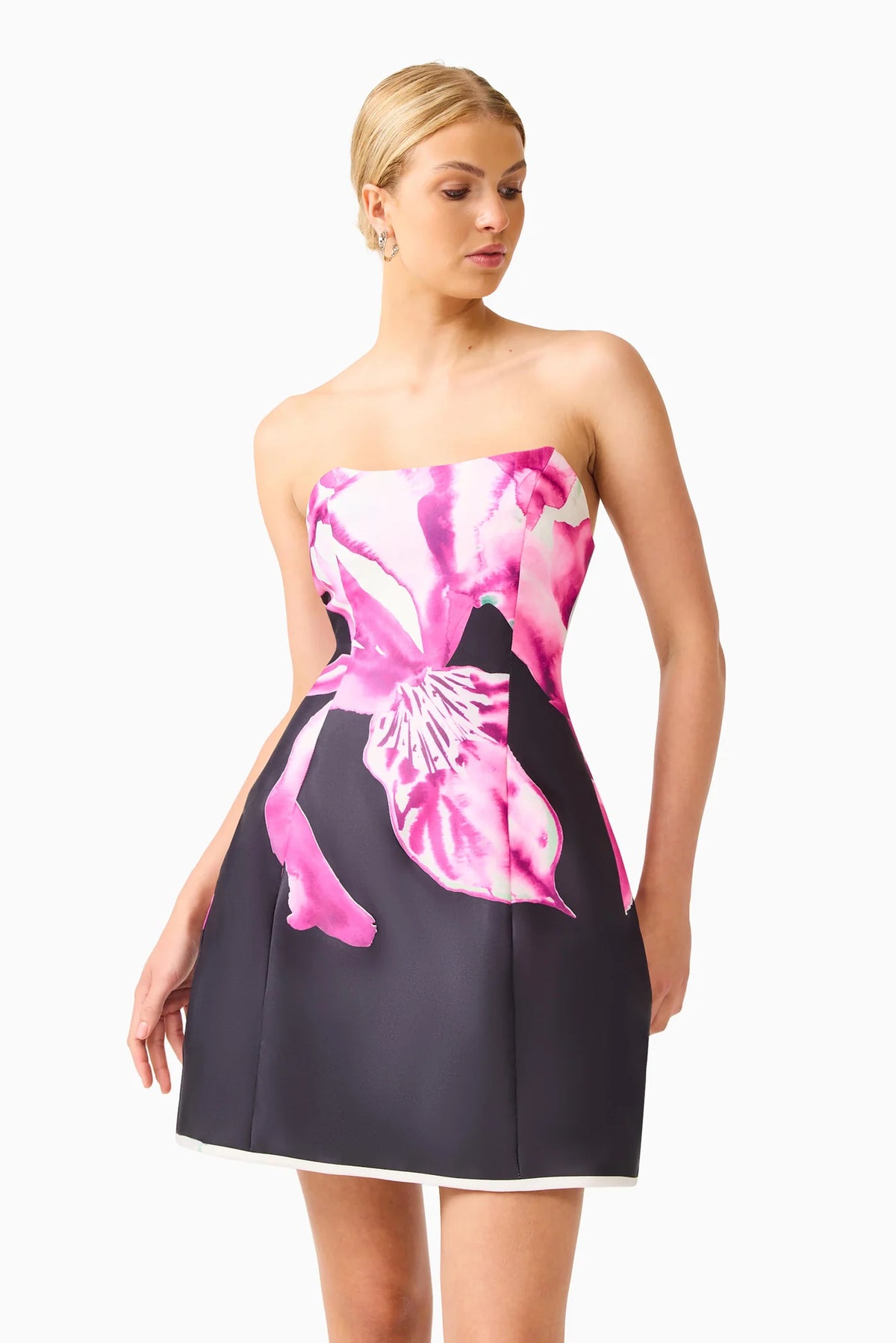 A woman modeling a strapless mini dress with a dark charcoal skirt and a vibrant pink and white floral print bodice, set against a plain white studio background.