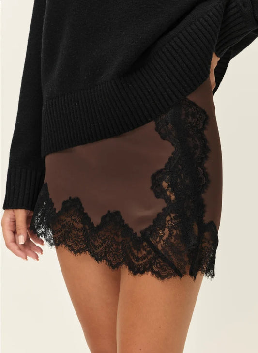 A close-up of a person wearing a black knit sweater and a sheer brown mini skirt with black lace trim along the hem and side edges, against a plain light background.