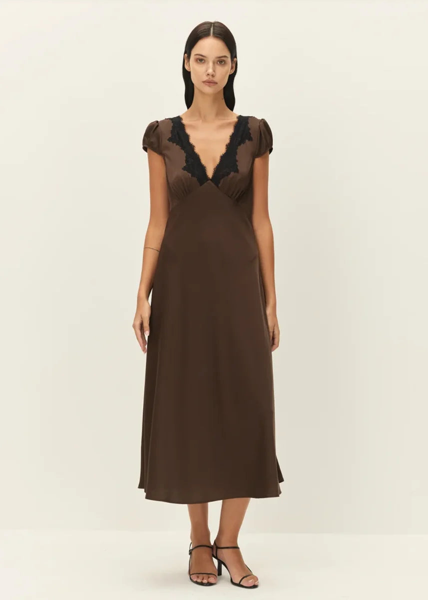A woman modeling a brown midi dress with a deep V-neckline, sheer cap sleeves, and black lace detailing at the neckline, set against a plain off-white studio background.