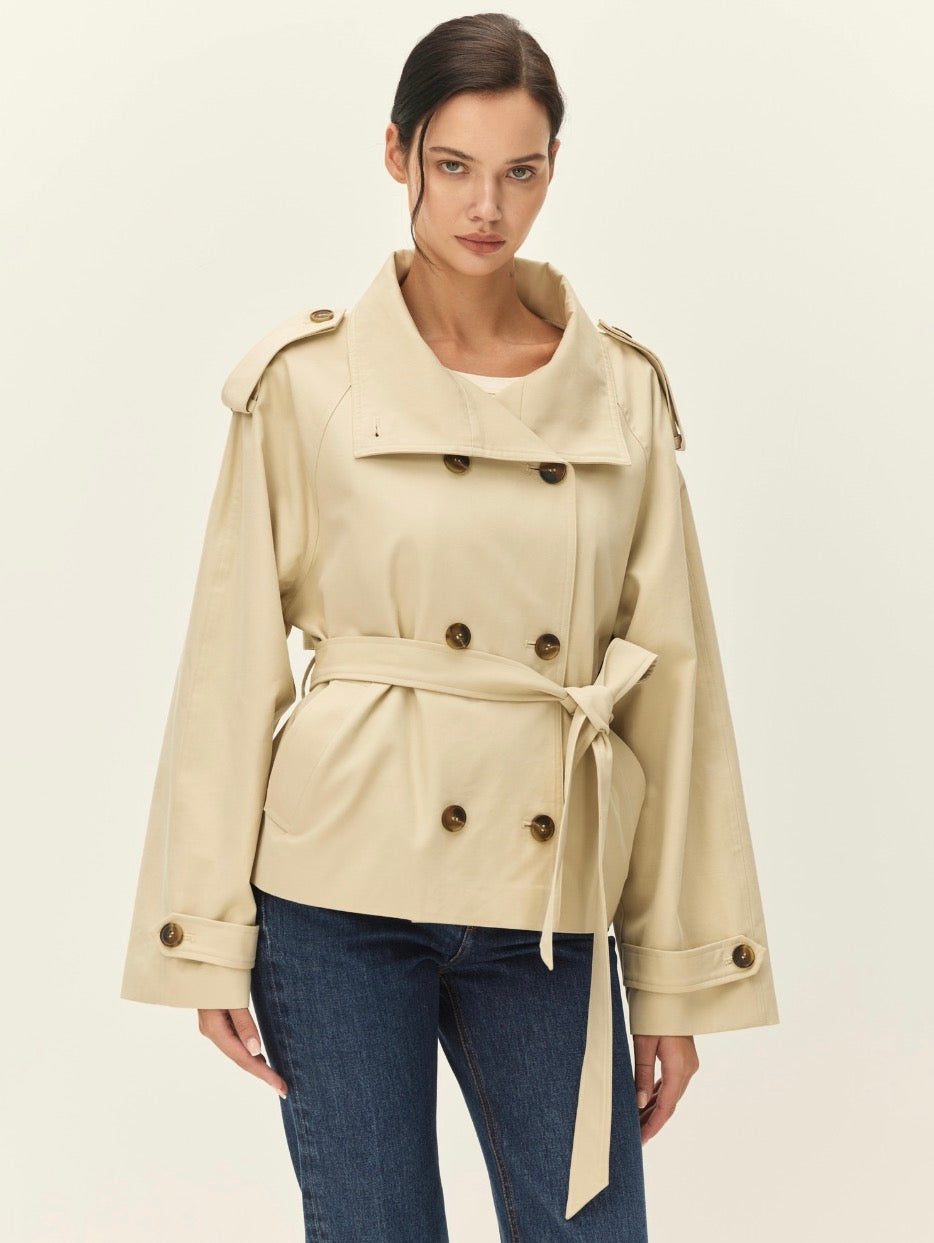A woman modeling a beige double-breasted trench coat with a tie waist belt, paired with dark blue jeans, against a plain off-white studio background.