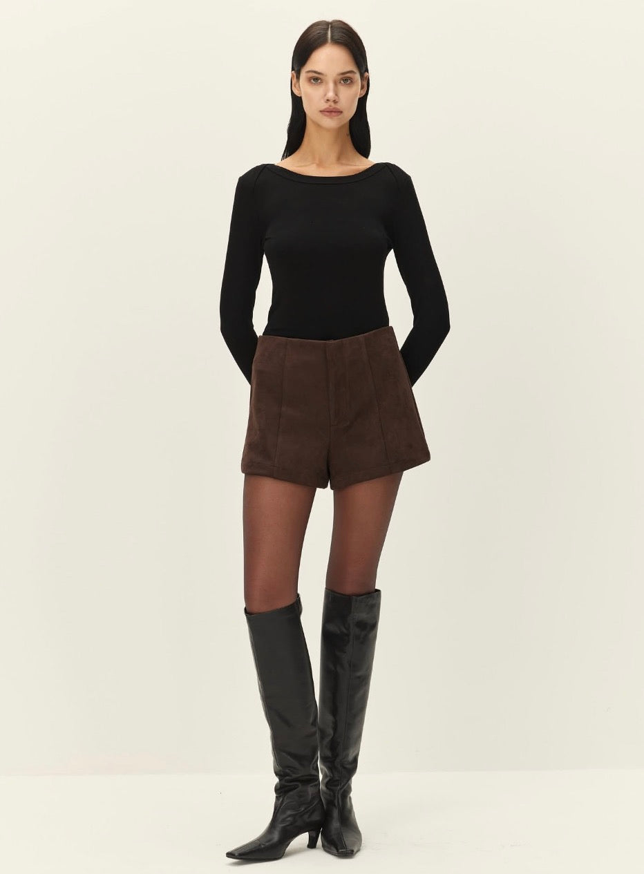 A model stands against a plain off-white studio background wearing a black long-sleeve boat-neck top, brown shorts, sheer black tights, and black knee-high boots.