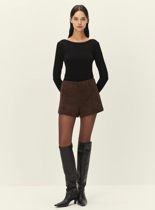 A model stands against a plain off-white studio background wearing a black long-sleeve boat-neck top, brown shorts, sheer black tights, and black knee-high boots.