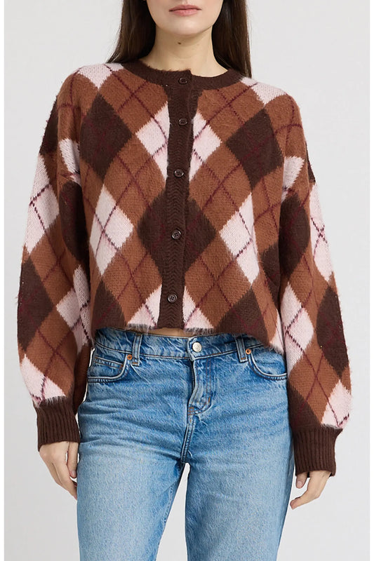 A woman wearing a cropped long-sleeve knit cardigan with a brown, pink, and white argyle pattern, paired with blue jeans, against a light gray studio background.