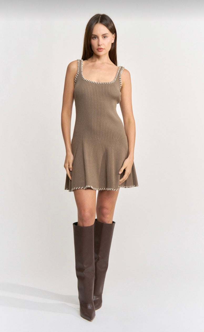 A woman modeling a sleeveless ribbed knit mini dress in taupe brown with silver beaded trim along the neckline and hem, paired with dark brown knee-high boots, against a light gray studio background.