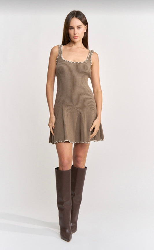 A woman modeling a sleeveless ribbed knit mini dress in taupe brown with silver beaded trim along the neckline and hem, paired with dark brown knee-high boots, against a light gray studio background.
