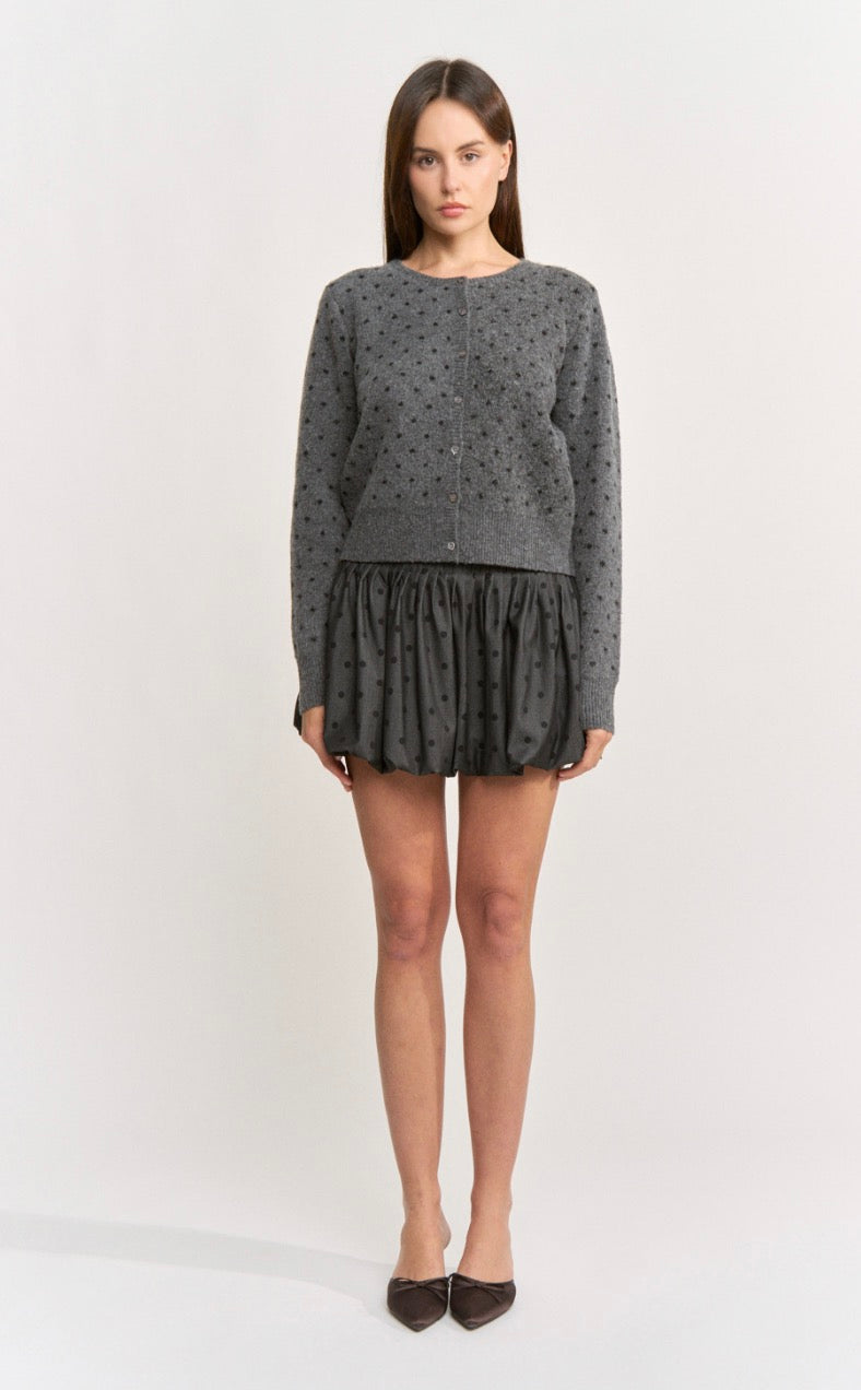 A woman modeling a dark gray long-sleeve cardigan with a small black polka dot pattern, paired with a matching dark gray ruffled mini skirt and dark pointed flats, against a plain light gray studio background.