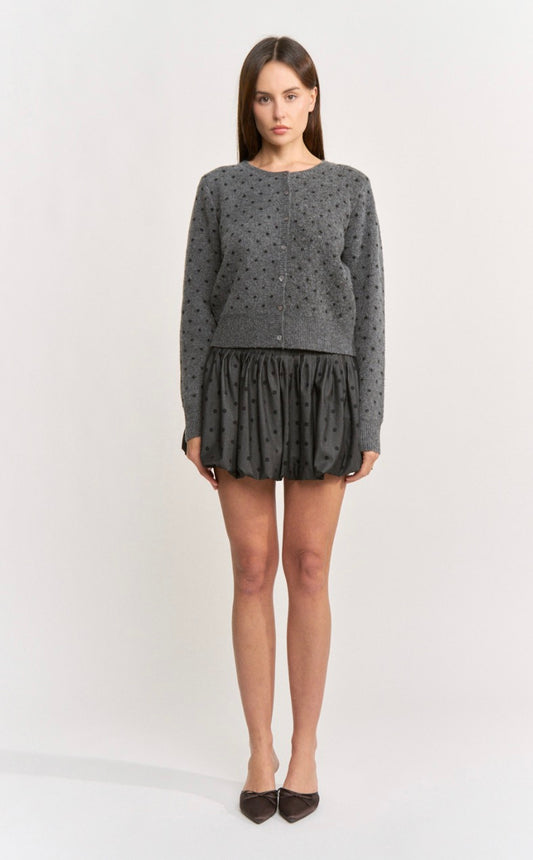 A woman modeling a dark gray long-sleeve cardigan with a small black polka dot pattern, paired with a matching dark gray ruffled mini skirt and dark pointed flats, against a plain light gray studio background.