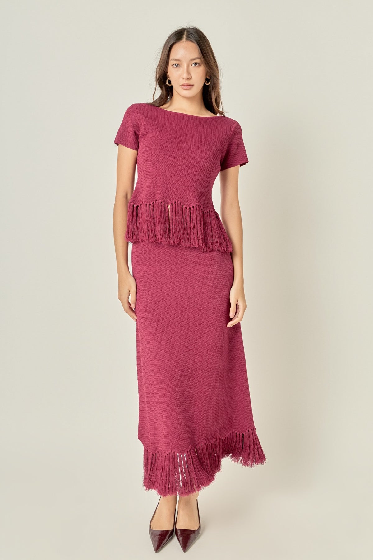 A woman modeling a dusty rose pink knit dress with short sleeves, a boat neckline, and fringe detailing at the peplum and hem, paired with dark brown pointed-toe heels against a light gray studio background.