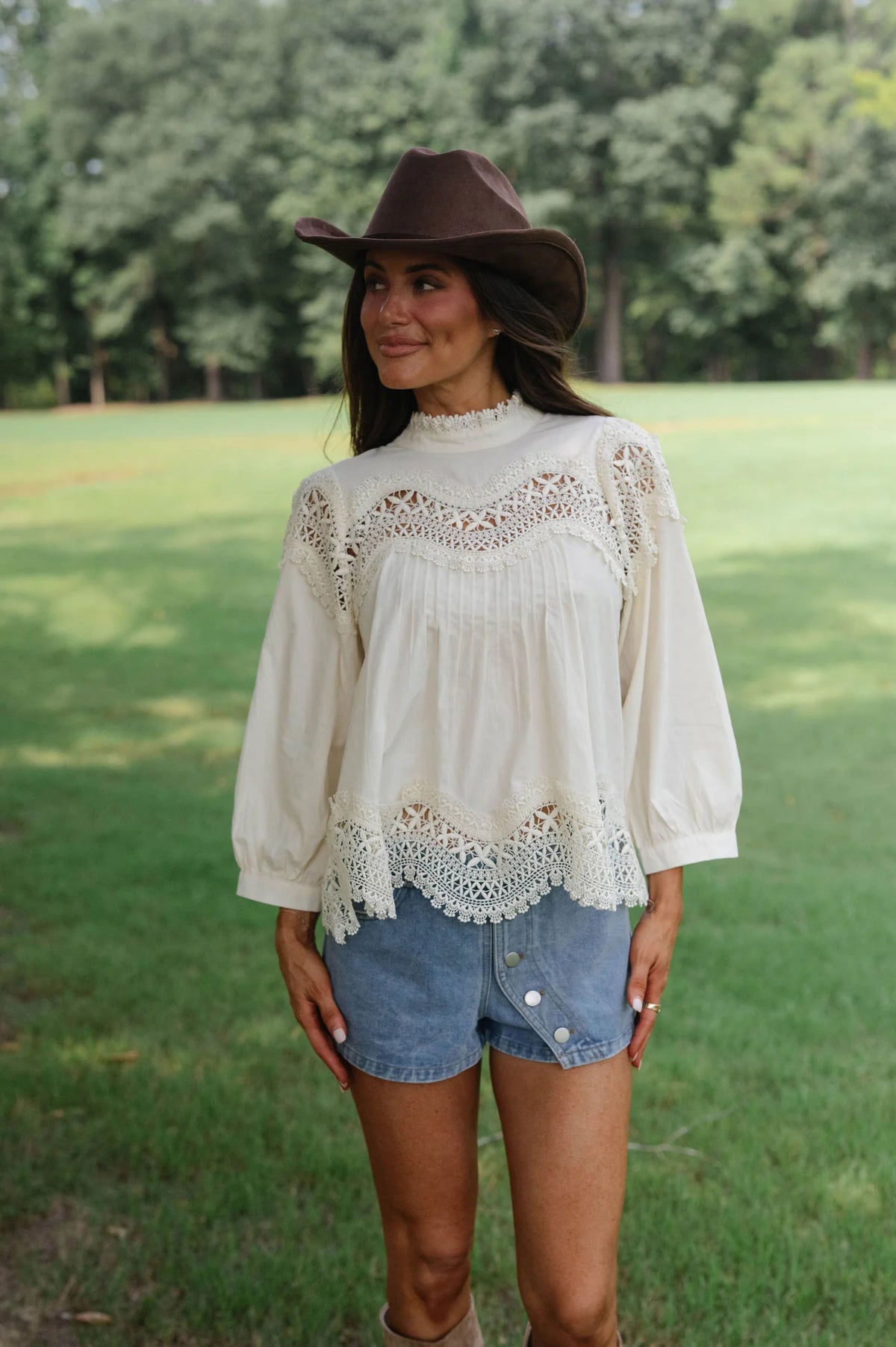 A woman standing outdoors in a grassy park wearing a cream bohemian-style lace blouse, denim shorts, and a brown cowboy hat.