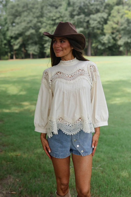 A woman standing outdoors in a grassy park wearing a cream bohemian-style lace blouse, denim shorts, and a brown cowboy hat.