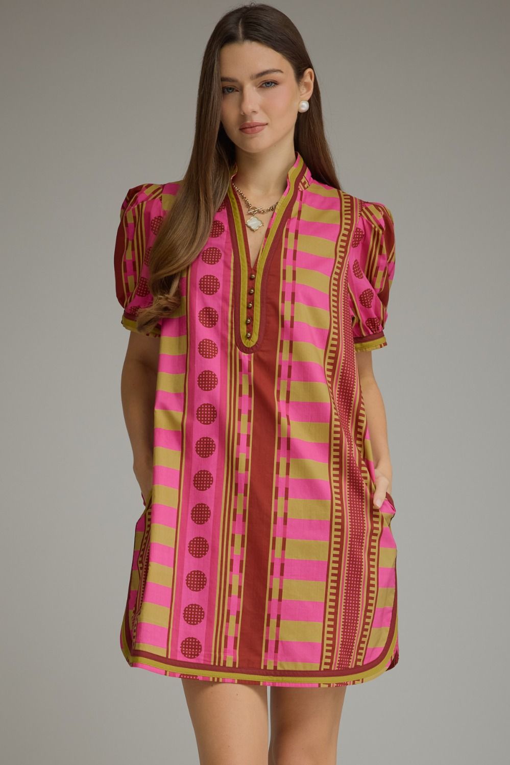 A woman modeling a short puff-sleeve tunic dress with vertical stripes in vibrant pink, olive green, and brown, featuring a V-neckline with gold trim and side pockets, set against a solid gray studio background.