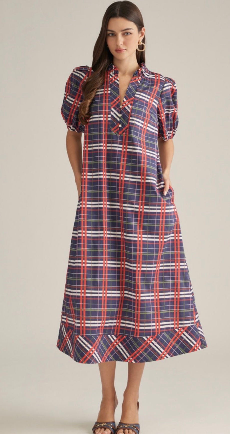 A woman modeling a midi-length plaid dress with puff sleeves, a tie neckline, and side pockets. The dress features a navy blue base with red and white plaid stripes, set against a solid light beige background.